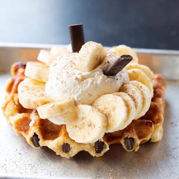 A golden-brown waffle topped with whipped cream, sliced bananas, and chocolate sticks.