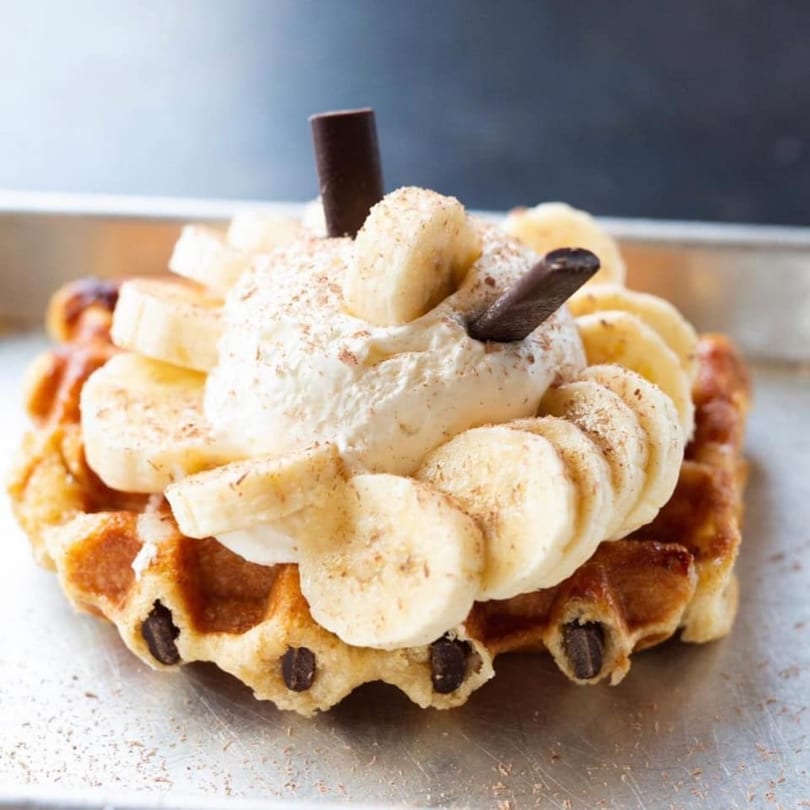 A golden-brown waffle topped with whipped cream, sliced bananas, and chocolate sticks.