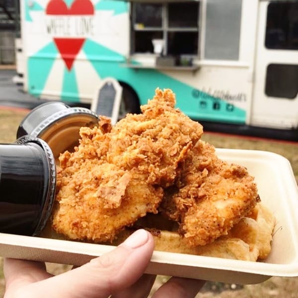 Hand holding a waffle and fried chicken dish with two sauce cups, in front of a food truck.