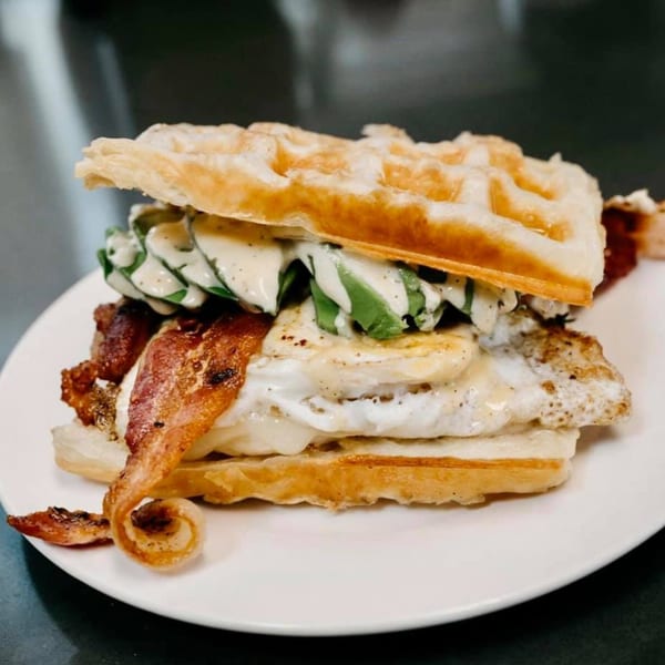 A waffle sandwich with bacon, fried egg, avocado, and creamy sauce on a white plate.