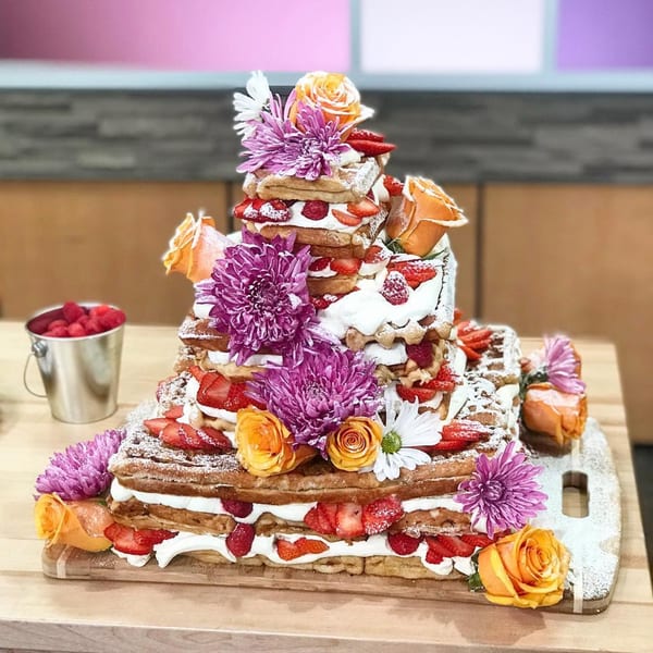 Tiered waffle cake with whipped cream, strawberries, raspberries, powdered sugar, and fresh flowers.