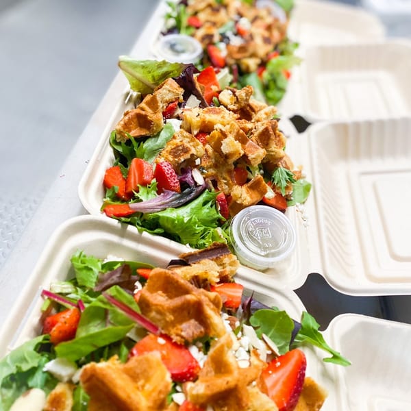 Multiple takeout containers filled with green salad, strawberries, waffle pieces, and a side of dressing.