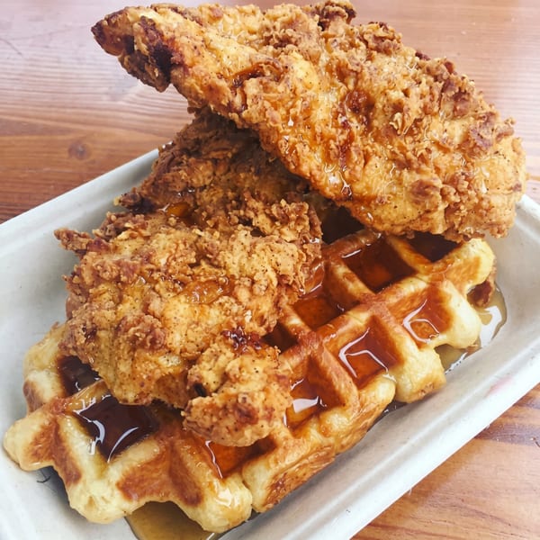 Crispy fried chicken pieces and a waffle drizzled with syrup in a white container on a wooden table.