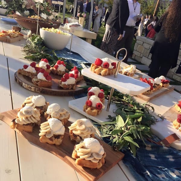 Waffles topped with whipped cream, raspberries, and bananas on a buffet table with people in the background.